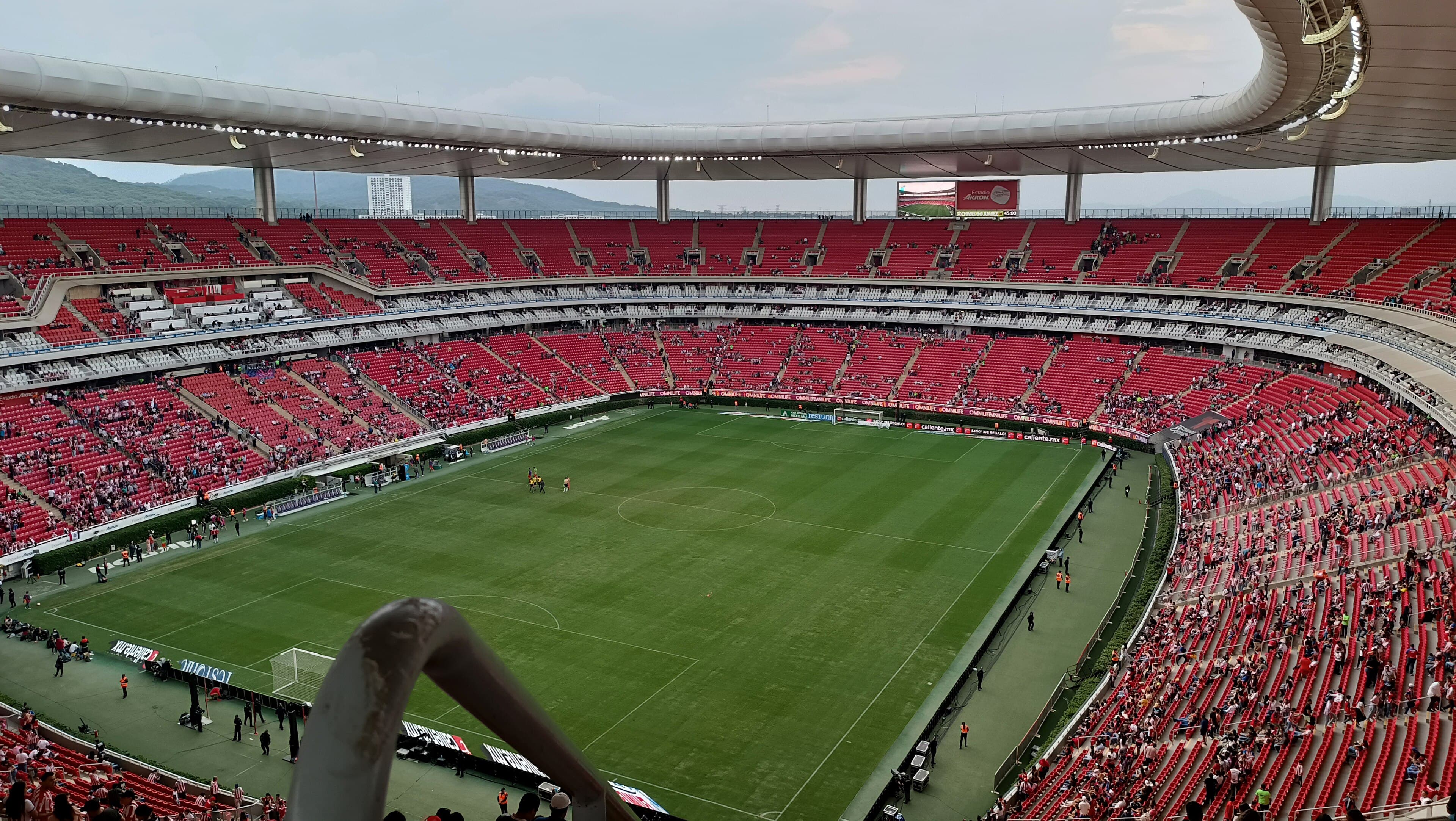 A wide interior view of Estadio Guadalajara with red seating, spectators, and the soccer pitch before a match.