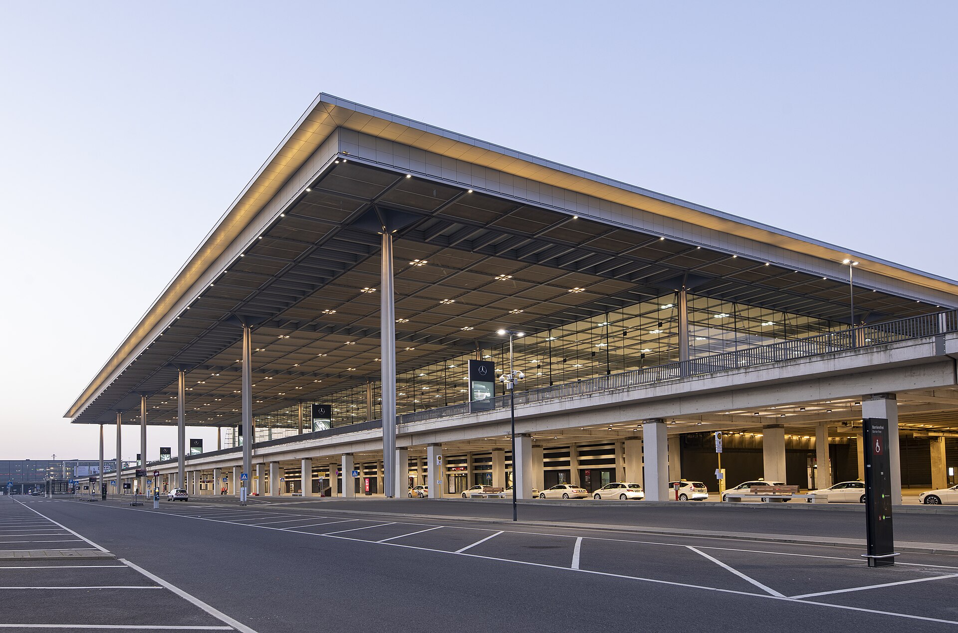 A large, modern airport terminal building is shown at dusk with its illuminated interior and expansive parking area in front — illustrative photograph